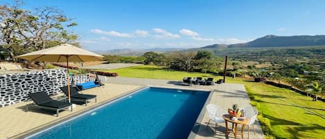 A rectangular swimming pool with a stone wall and lounge chairs. There is a table with a basket of fruit and a bottle. The pool is surrounded by a paved area with a view of distant hills and a clear sky.
