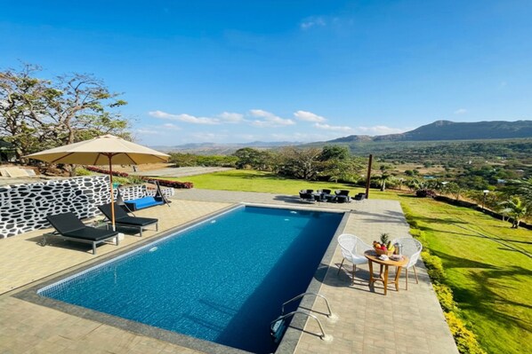 A rectangular swimming pool with a stone wall and lounge chairs. There is a table with a basket of fruit and a bottle. The pool is surrounded by a paved area with a view of distant hills and a clear sky. - Mayank Farm - Valley View Pool Villa (Mawal)