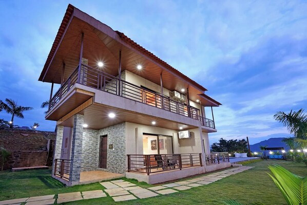 A two-story house with a balcony on the upper floor, stone walls, and a well-lit outdoor area.