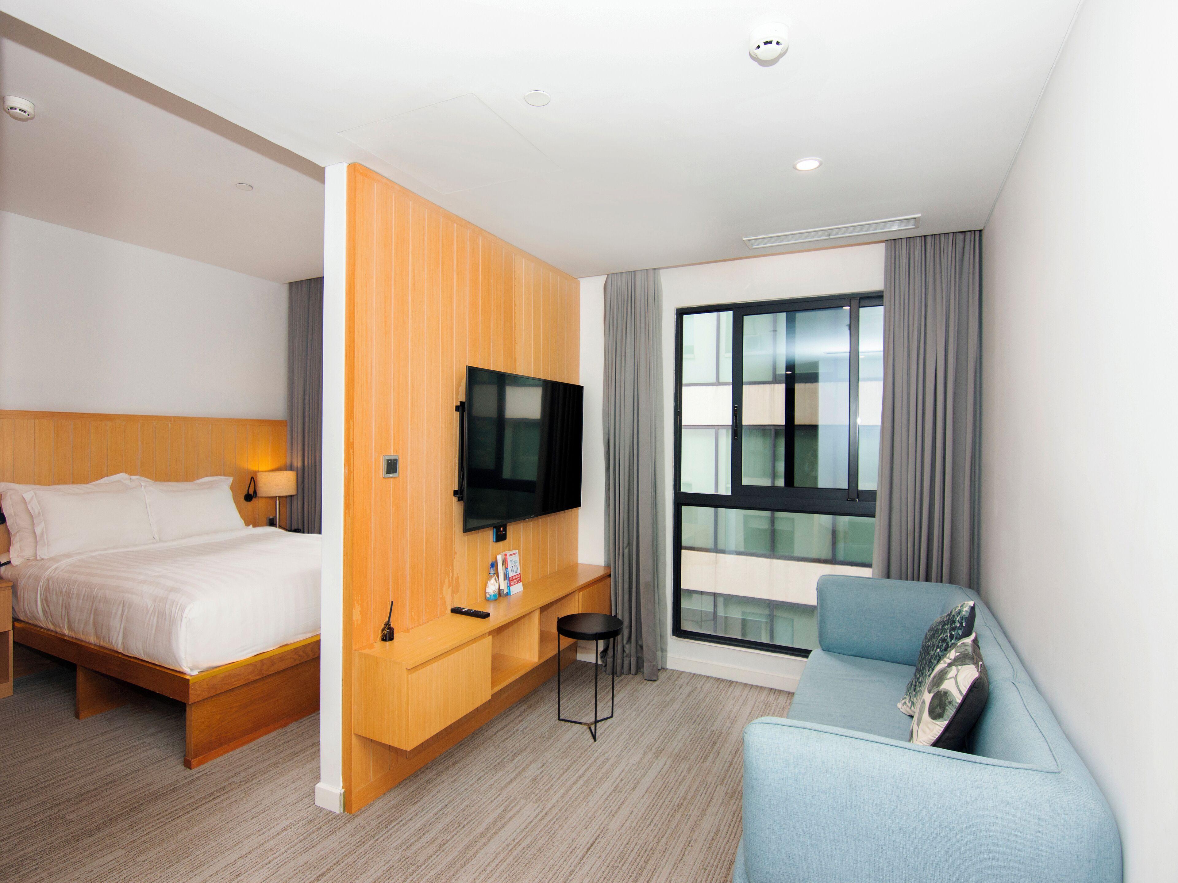 Une chambre d’hôtel avec un lit, un téléviseur fixé sur un panneau en bois, une petite table, une chaise et une grande fenêtre avec des rideaux.