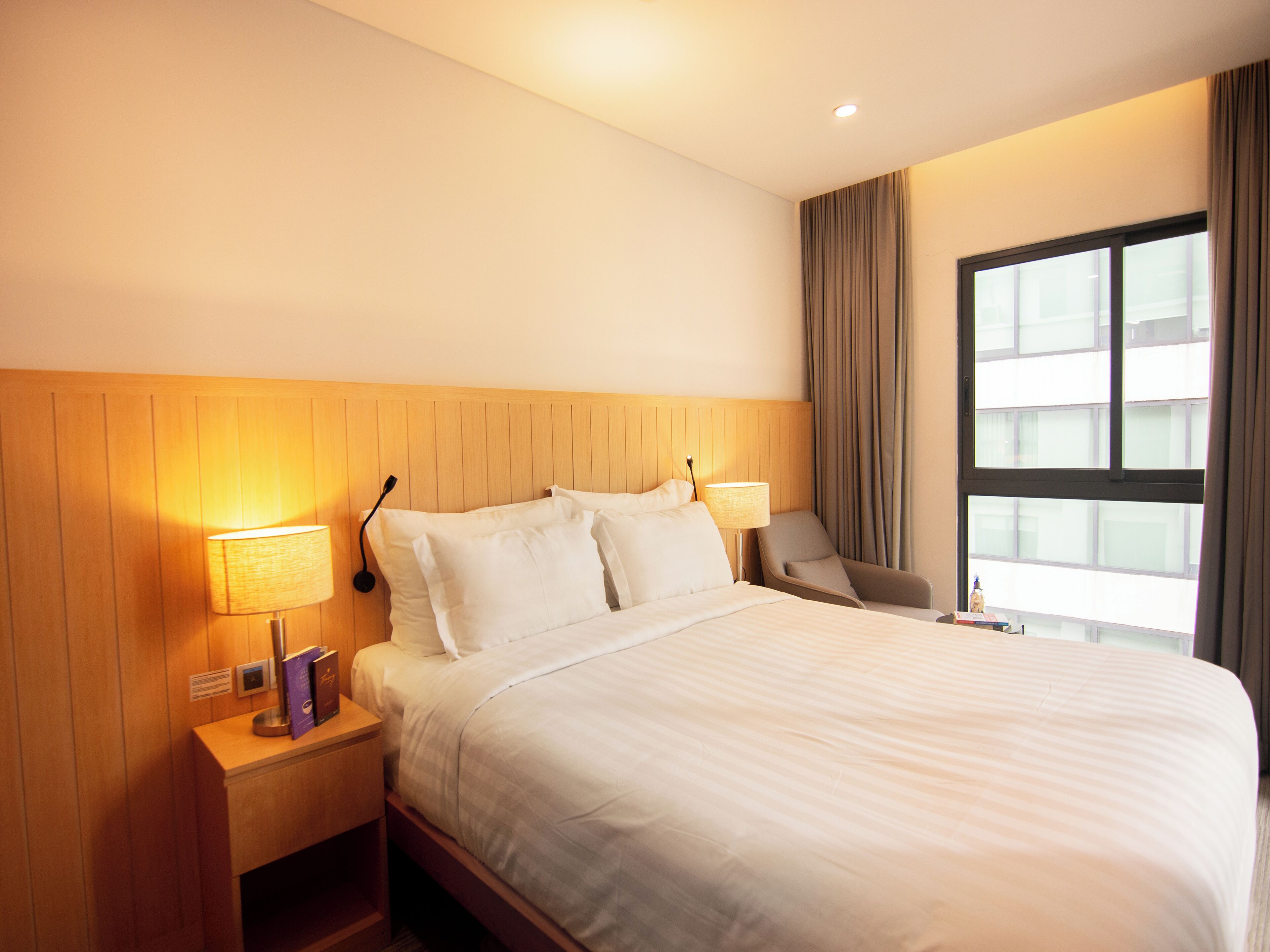 Habitación de hotel con una cama grande, dos mesitas de noche, una silla y una ventana con cortinas.