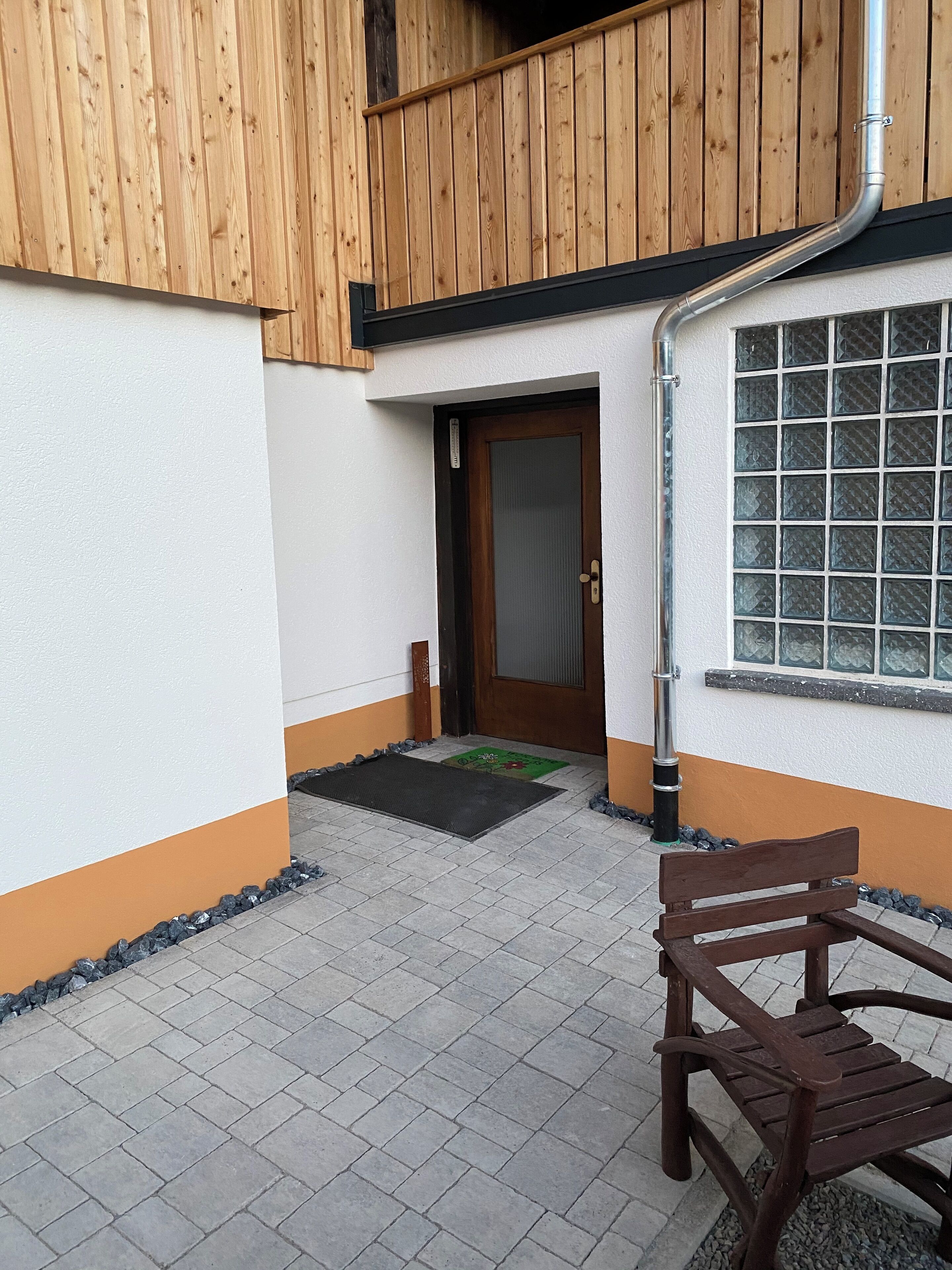 A wooden exterior wall with a door and window, a bench, and a tiled patio area.