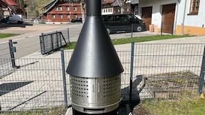 Uma churrasqueira portátil para áreas externas com chaminé cônica, posicionada em área pavimentada, com cerca de metal ao fundo.