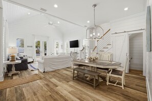 A spacious room with a wooden floor, a dining table with chairs, a bench, a ceiling fan, and a TV mounted on the wall. - Sandy Toes on Parks Edge (Orange Beach)