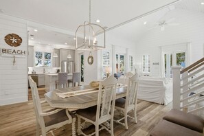 A spacious dining area with a wooden table and white chairs, a chandelier, and a view of the kitchen and living area in the background. - Sandy Toes on Parks Edge (Orange Beach)