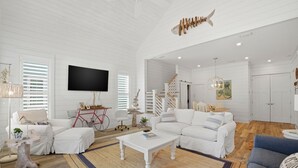 Un salón amplio con muebles blancos, un televisor de pantalla plana y una escalera que lleva a otro nivel.
