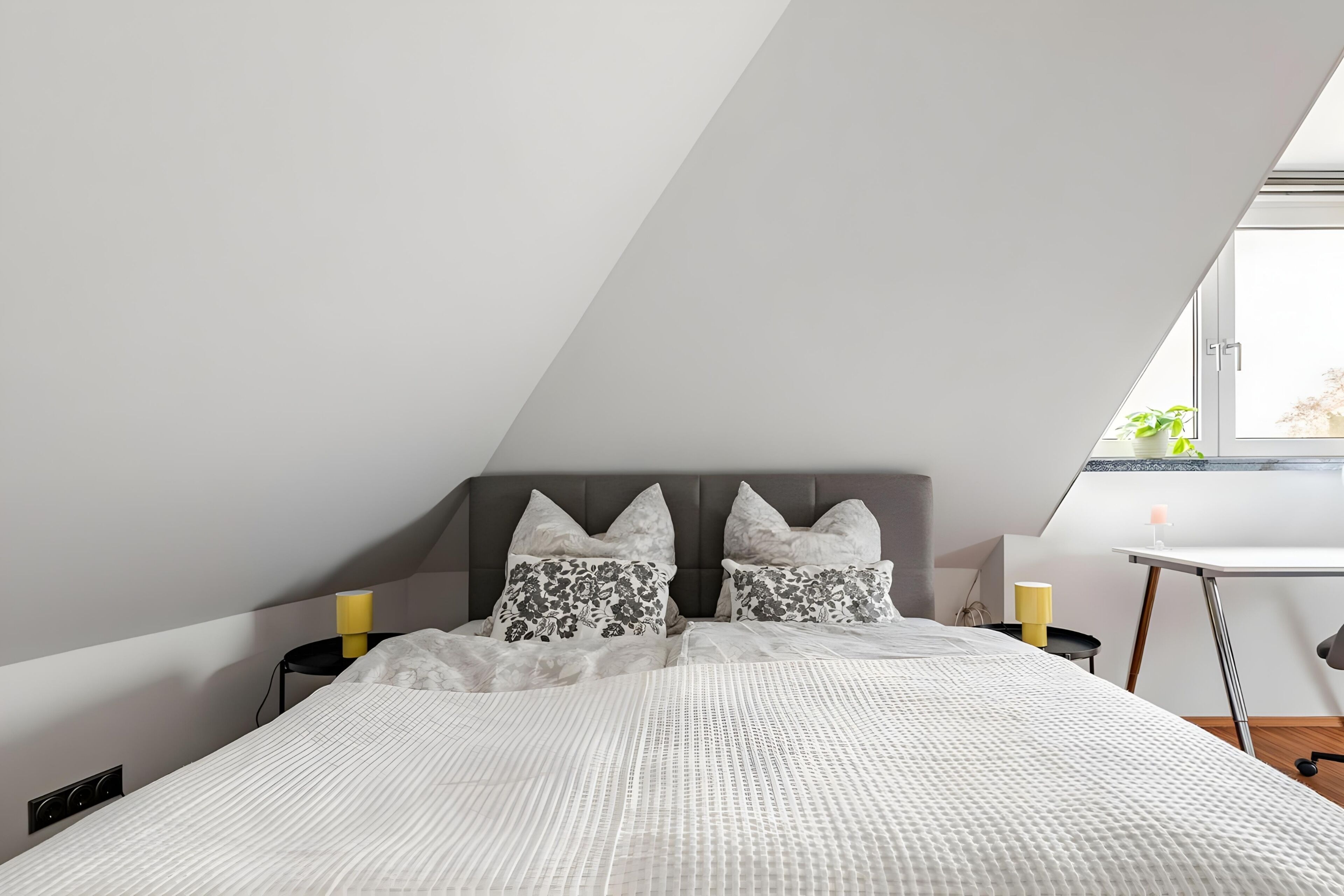 A modern bedroom with a sloped ceiling, a bed with a white quilt, two decorative pillows, a bedside table with a yellow candle, and a small side table with a candle.