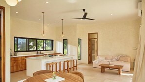 Una cocina moderna con una isla central, mesa de comedor de madera y un sofá en una sala de estar luminosa con ventilador de techo.
