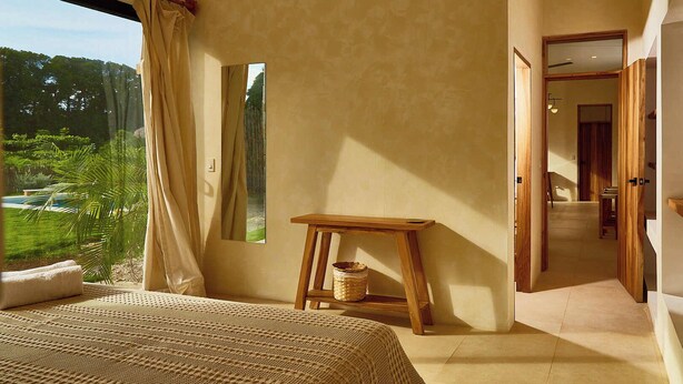 A bedroom with a large bed, a small wooden table, a mirror, and a view of a garden.