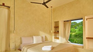 Un dormitorio con cama, ventilador de techo, ventana con vista a follaje y mesita de noche con un florero.