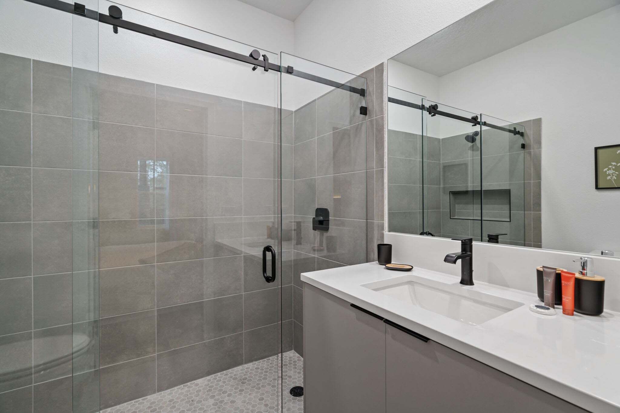 Un baño moderno con ducha de vidrio, un espejo grande y una encimera blanca con artículos de aseo.