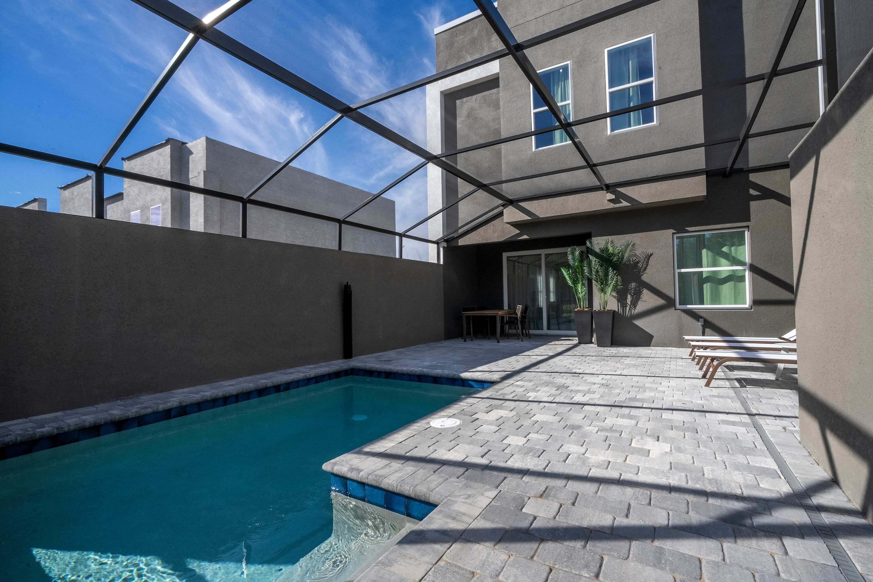 Una piscina con borde de baldosas, un patio cubierto y un edificio con amplias ventanas y techo plano.