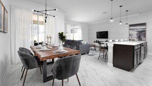 Eine moderne Küche mit Kochinsel, ein Essbereich mit Holztisch und grauen Stühlen sowie eine Wohnbereich mit Sofa und Fernseher.