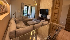 Un saló modern amb un sofà modular, una taula de centre de vidre, un televisor de pantalla plana fixat a una paret amb relleu de maó i una porta corredissa de vidre que dona a un pati exterior.
