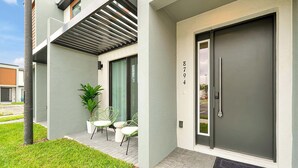 Um edifício moderno com uma porta cinza identificada com o número "8794", uma pequena área de estar ao ar livre com cadeiras e uma planta, e um gramado bem cuidado.