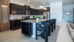 A modern kitchen with dark cabinetry, a central island with bar stools, and stainless steel appliances. - Last Minute Disney Getaway! Luxury Villa with Private Pool, Game Room & Themed Bedrooms. Book Now! #159555 (Orlando)