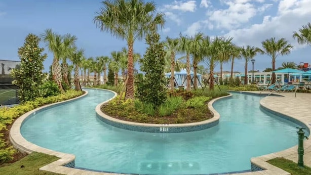 A curved swimming pool surrounded by palm trees and greenery, with lounge chairs and a covered area in the background. - Last Minute Disney Getaway! Luxury Villa with Private Pool, Game Room & Themed Bedrooms. Book Now! #159555 (Orlando)