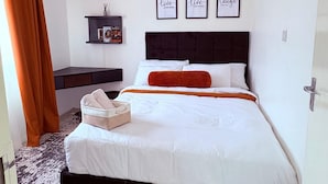 Een netjes opgemaakt bed met witte lakens en een oranje plaid, een nachtkastje met een lamp, en drie motiverende posters aan de muur.