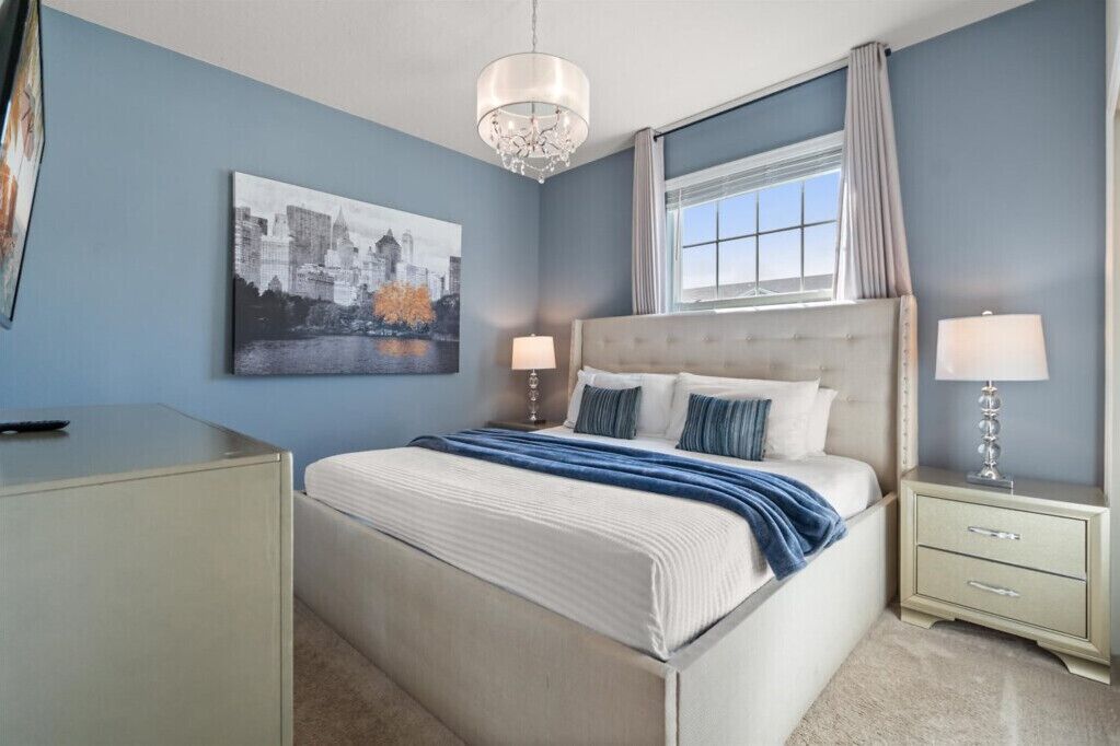 Une chambre à coucher avec un lit, des tables de chevet, un lustre, une fenêtre avec des rideaux et un tableau représentant un paysage urbain accroché au mur.
