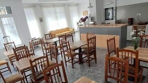 A spacious dining area with wooden tables and chairs, a kitchen in the background, and large windows allowing natural light.