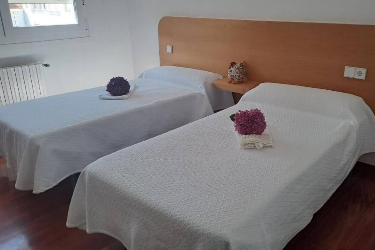 Dos camas individuales con sábanas blancas, un cabecero de madera, una mesita de noche con un adorno y un radiador al fondo.
