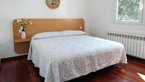 A bedroom with a wooden headboard, a white bedspread, a bedside table with a vase of flowers, a round mirror on the wall, and a window with a view of trees.