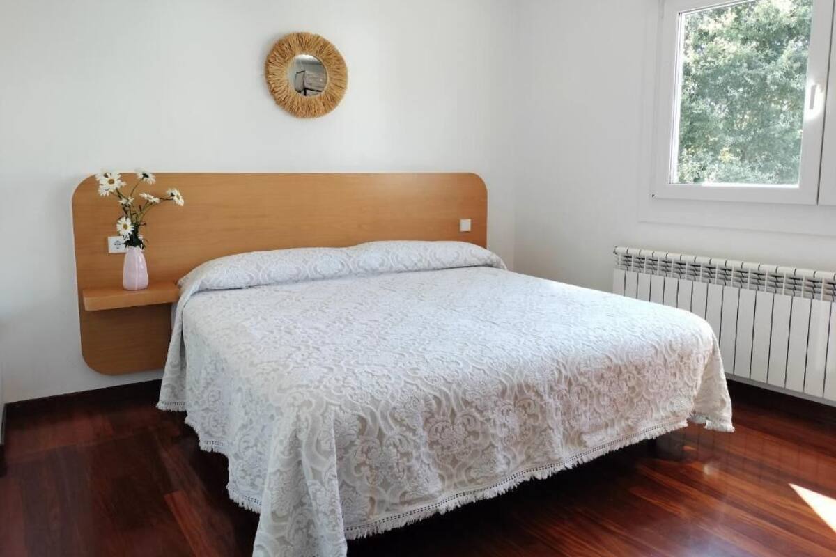 Un dormitorio con cabecera de madera, colcha blanca, mesita de noche con un florero, espejo redondo en la pared y ventana con vista a árboles.