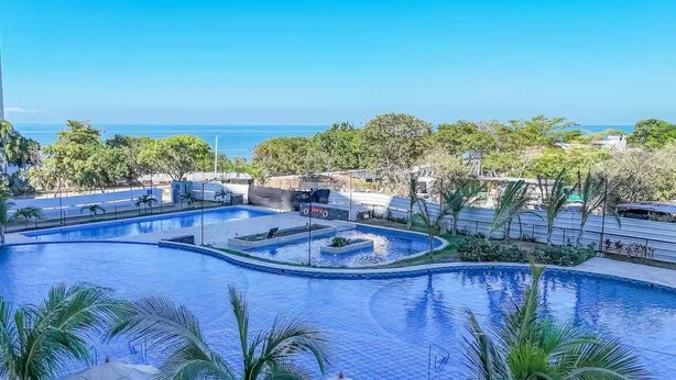 A large, curved swimming pool with a view of the ocean, surrounded by palm trees and a clear blue sky. - Cozy 1-bedroom apartment in charming Urb Don Jaca with AC (Urb Don Jaca)