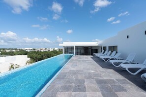 Une piscine sur le toit, avec une eau d’un bleu cristallin, entourée de chaises longues blanches et bordée d’un bâtiment moderne aux grandes fenêtres.