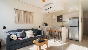 Ein modernes Wohnzimmer mit grauem Sofa, einem Holzschemel und einer Küchenzeile inklusive Kühlschrank, Mikrowelle und Barhocker.