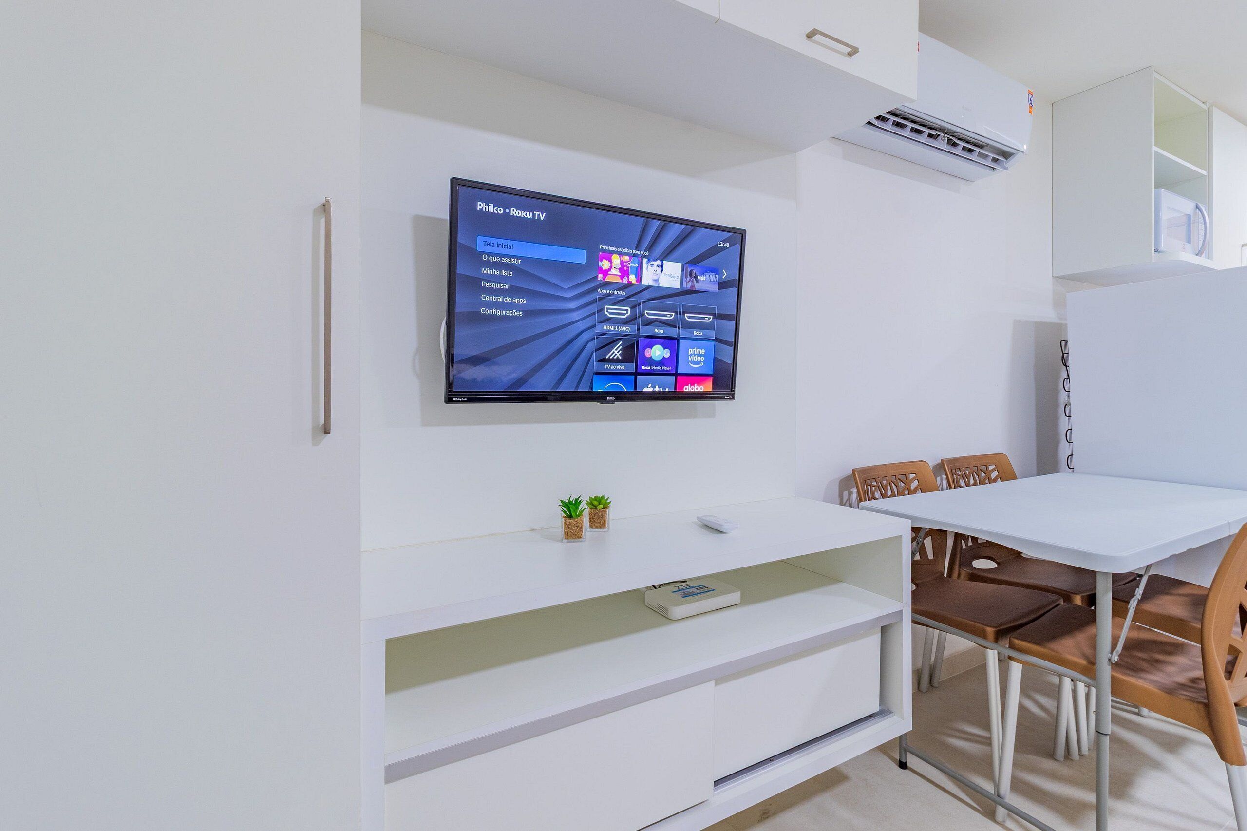 A modern living room with a flat-screen TV mounted on the wall, a white entertainment unit below, and a dining area with a table and chairs.