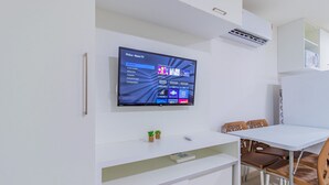 A modern living room with a flat-screen TV mounted on the wall, a white entertainment unit below, and a dining area with a table and chairs. - Beach Way Carneiros #209 - Quadruple by Carpediem (Tamandaré)