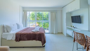 A modern hotel room with a bed, a TV, a desk, and a view of greenery outside. - Beach Way Carneiros #209 - Quadruple by Carpediem (Tamandaré)