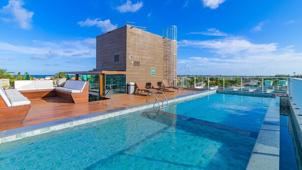 A rooftop infinity pool with wooden deck and lounge chairs, a modern building with a glass facade, and a clear blue sky. - Beach Way Carneiros #209 - Quadruple by Carpediem (Tamandaré)