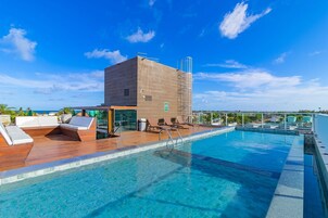 Una piscina infinita en la azotea con terraza de madera y sillones de descanso, un edificio moderno con fachada de vidrio y un cielo azul despejado.