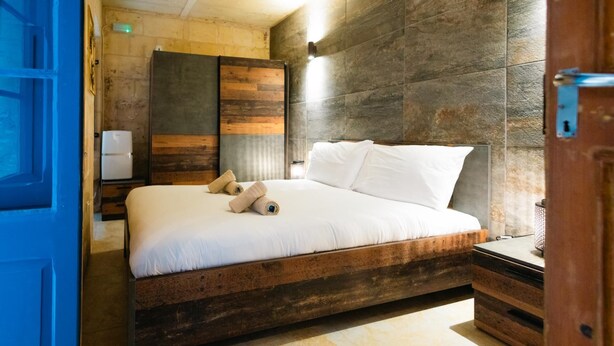 A modern bedroom with a wooden bed frame, white bedding, and a stone wall. - Comfy 2 BR Flat in the Heart of Floriana (Floriana)