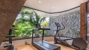 Un gimnasio moderno con cinta de correr, elíptica y bicicleta estática, que presenta una pared de piedra y ventanas amplias con vistas a zonas verdes.