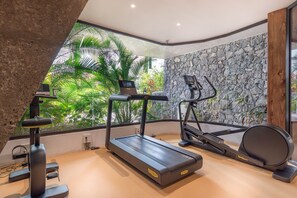 A modern gym with a treadmill, elliptical, and stationary bike, featuring a stone wall and large windows with a tropical view. - JARDINES - ISORA 0.3 GARDEN VIEW (Arona)