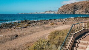 Un paisaje costero con una playa rocosa, una escalera que desciende hacia la orilla y un skyline distante.