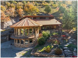 Una casa in pietra con una veranda in legno e un tetto in metallo, circondata da un giardino e un pendio boscoso.