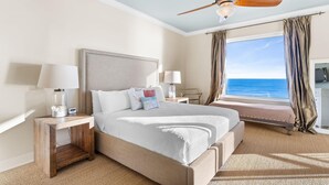 A hotel room with a bed, two bedside tables with lamps, a ceiling fan, a view of the ocean, and a window with curtains. - Indigo East 1402 (Pensacola)