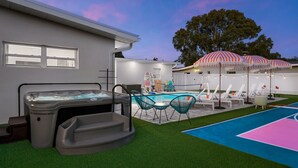 Un patio trasero con un jacuzzi, una piscina, sillas de descanso y una zona de estar cubierta con sombrillas.