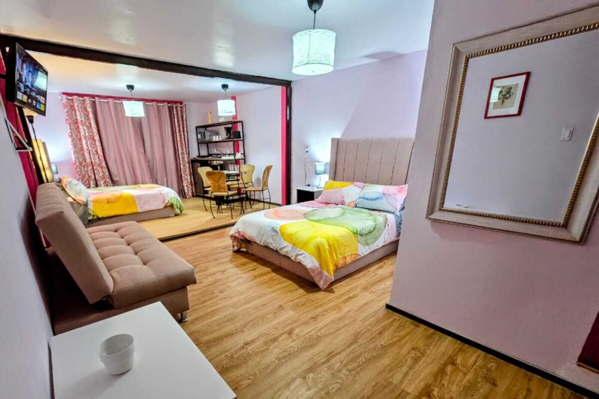 A hotel room with a sofa, a bed with colorful bedding, a small table with a cup, and a dining area with chairs and a table.