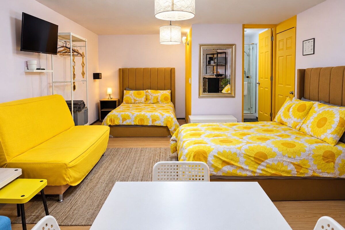 A hotel room with a yellow sofa, two beds with floral bedding, a white table, and a TV mounted on the wall.