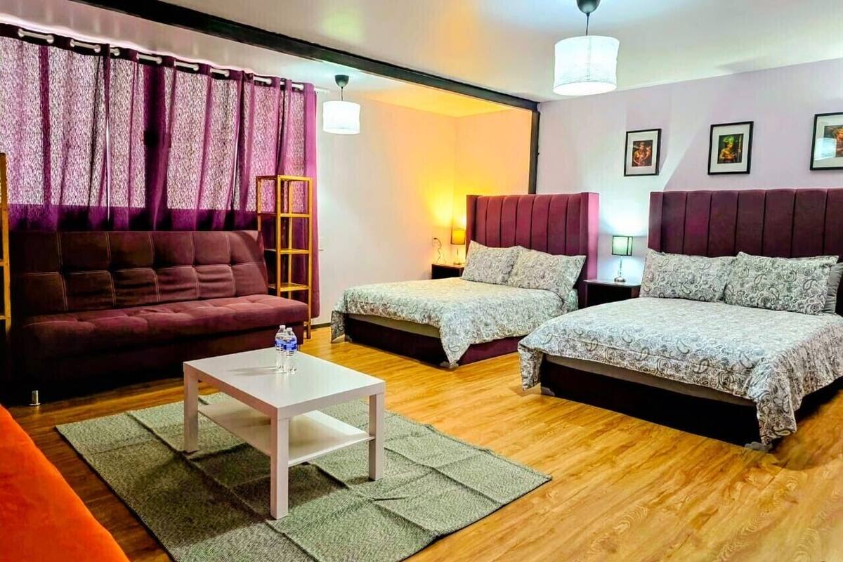A room with a sofa, two beds, a coffee table, and a lamp.