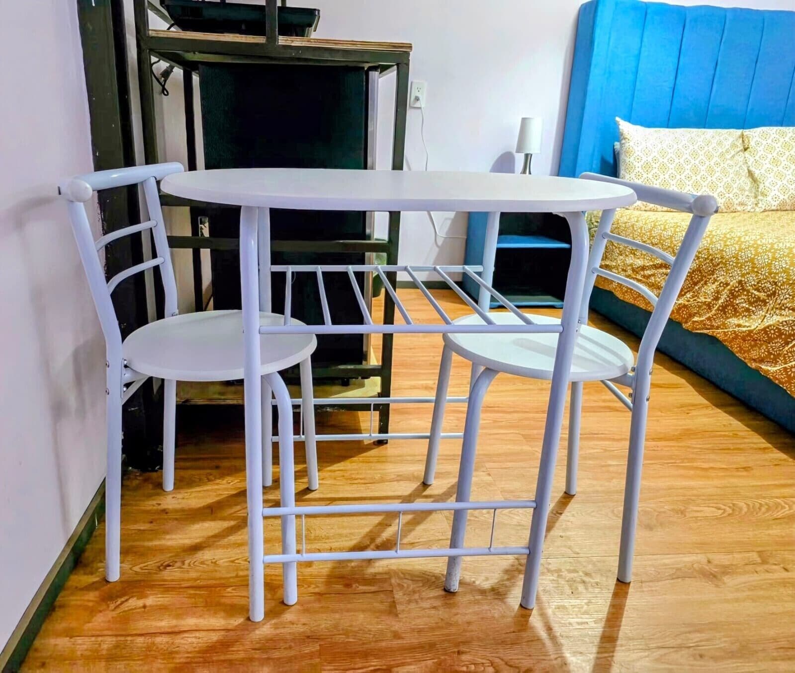 Una pequeña mesa blanca con estructura metálica y dos sillas en una habitación con piso de madera, un cabecero azul y una colcha estampada.