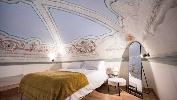 Un dormitorio con una cama, un taburete pequeño, un espejo y un techo con diseños decorativos.