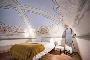 Een slaapkamer met een bed, een klein krukje, een spiegel en een plafond met decoratieve patronen.