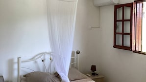 Quarto com cama, ventilador de teto, mesa de cabeceira, mesa lateral e janela com moldura de madeira.