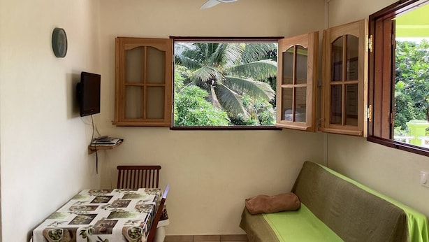 Um quarto pequeno com ventilador de teto, janela com vista para palmeiras, mesa com toalha de mesa estampada e sofá verde.